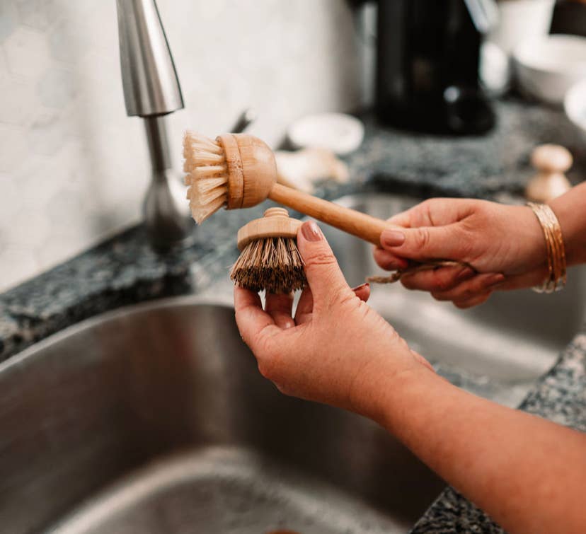 Two wooden dish brushes held by hands in front of a kitchen sink.