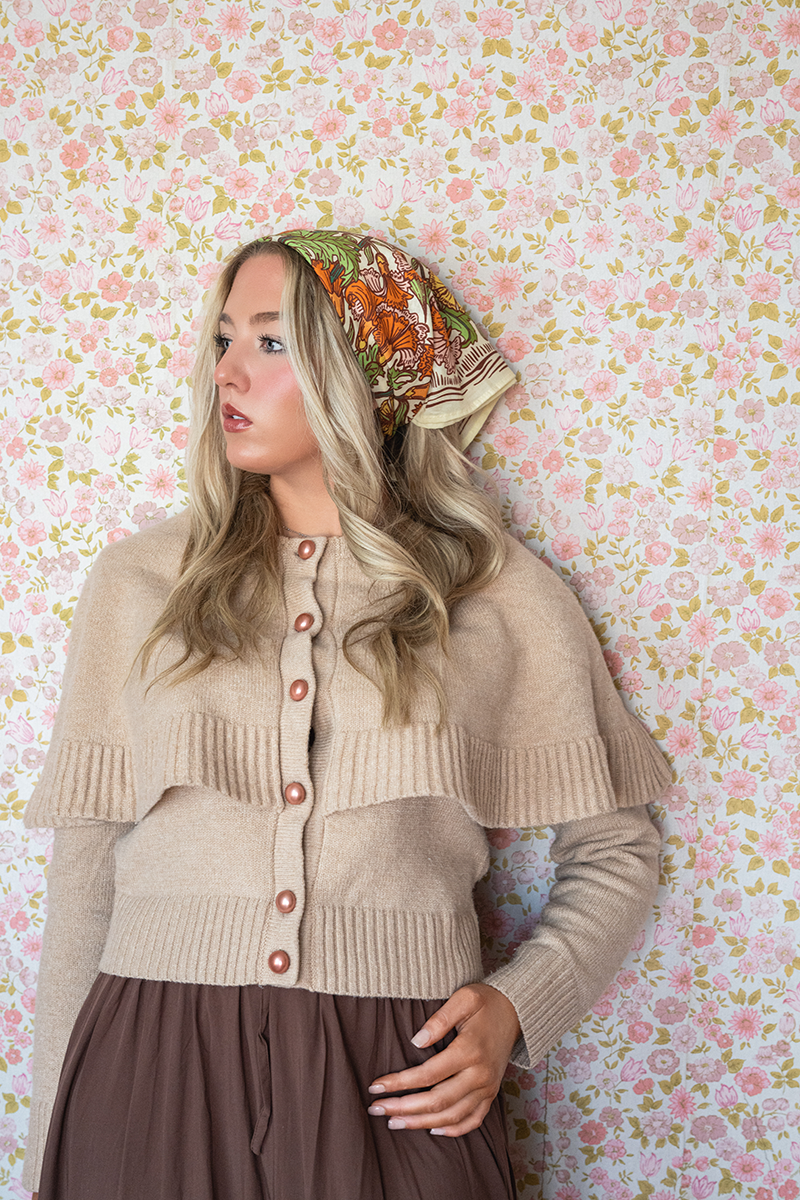 Woman wearing a beige cardigan and patterned headscarf against a floral wallpaper background