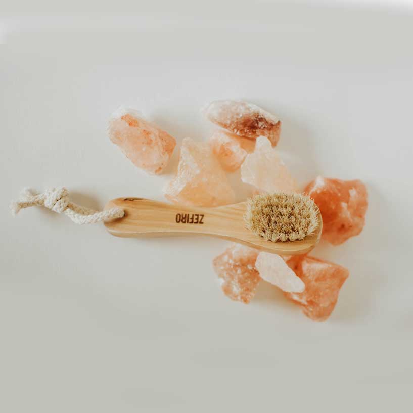 Bath brush with a wooden handle and natural bristles, surrounded by pink Himalayan salt crystals on a light background.