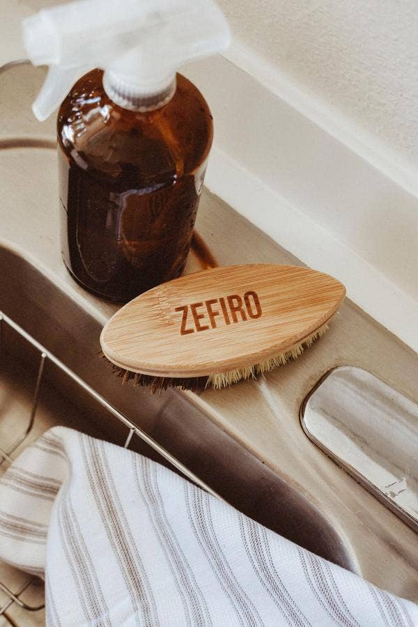 Bottle of brown liquid with a white spray nozzle, wooden brush labeled 'Zefiro', and striped towel on a beige surface.