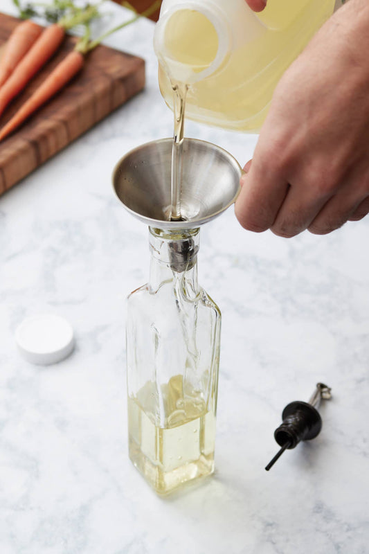 Person pouring liquid into a small funnel over a bottle on a marble surface.