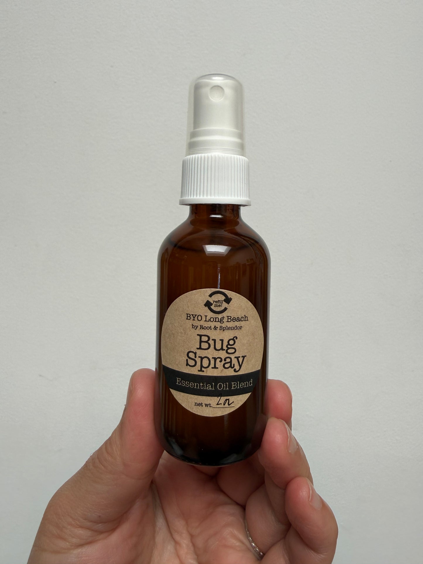 A hand holding a small amber glass spray bottle labeled 'Bug Spray' against a plain light gray background.