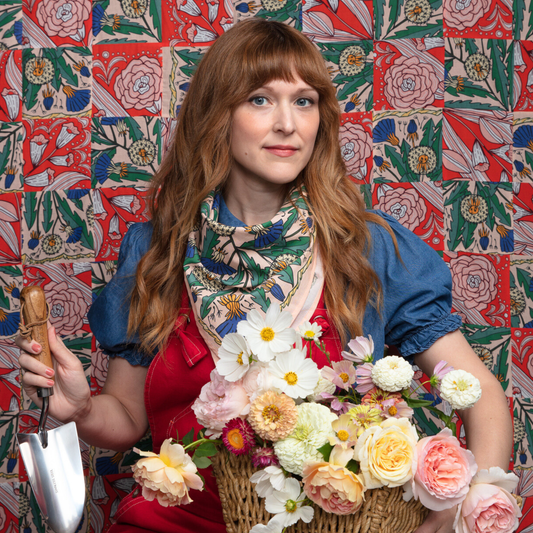 Woman holding a basket of flowers and a gardening tool against a colorful floral-patterned wall.