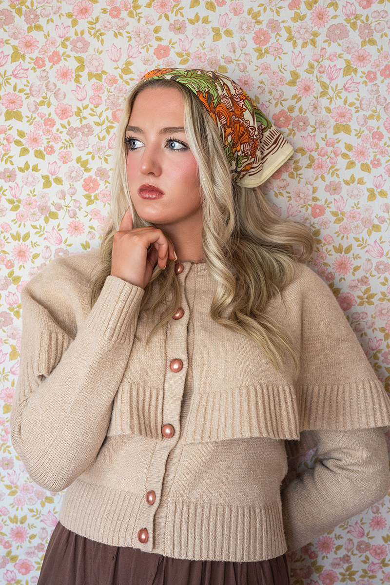 Woman wearing a beige cardigan and colorful headscarf against a floral wallpaper background