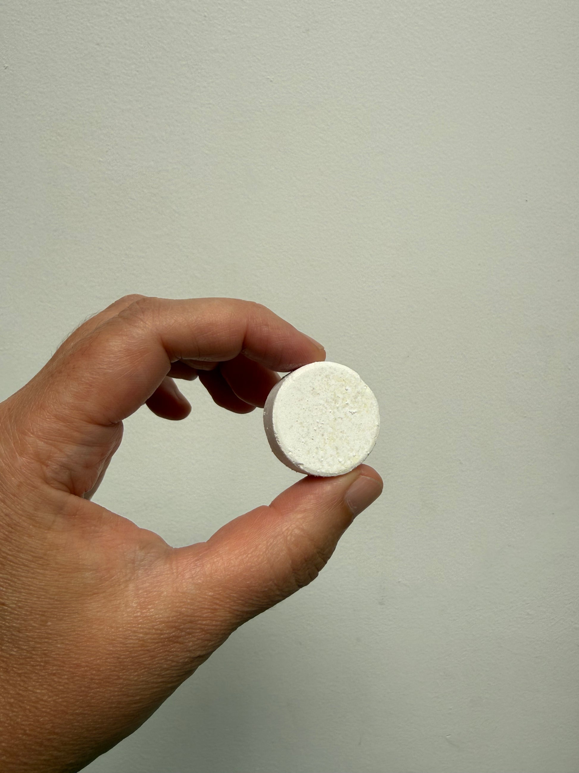 A hand holding a small, round, off-white tablet against a plain, light gray wall background.