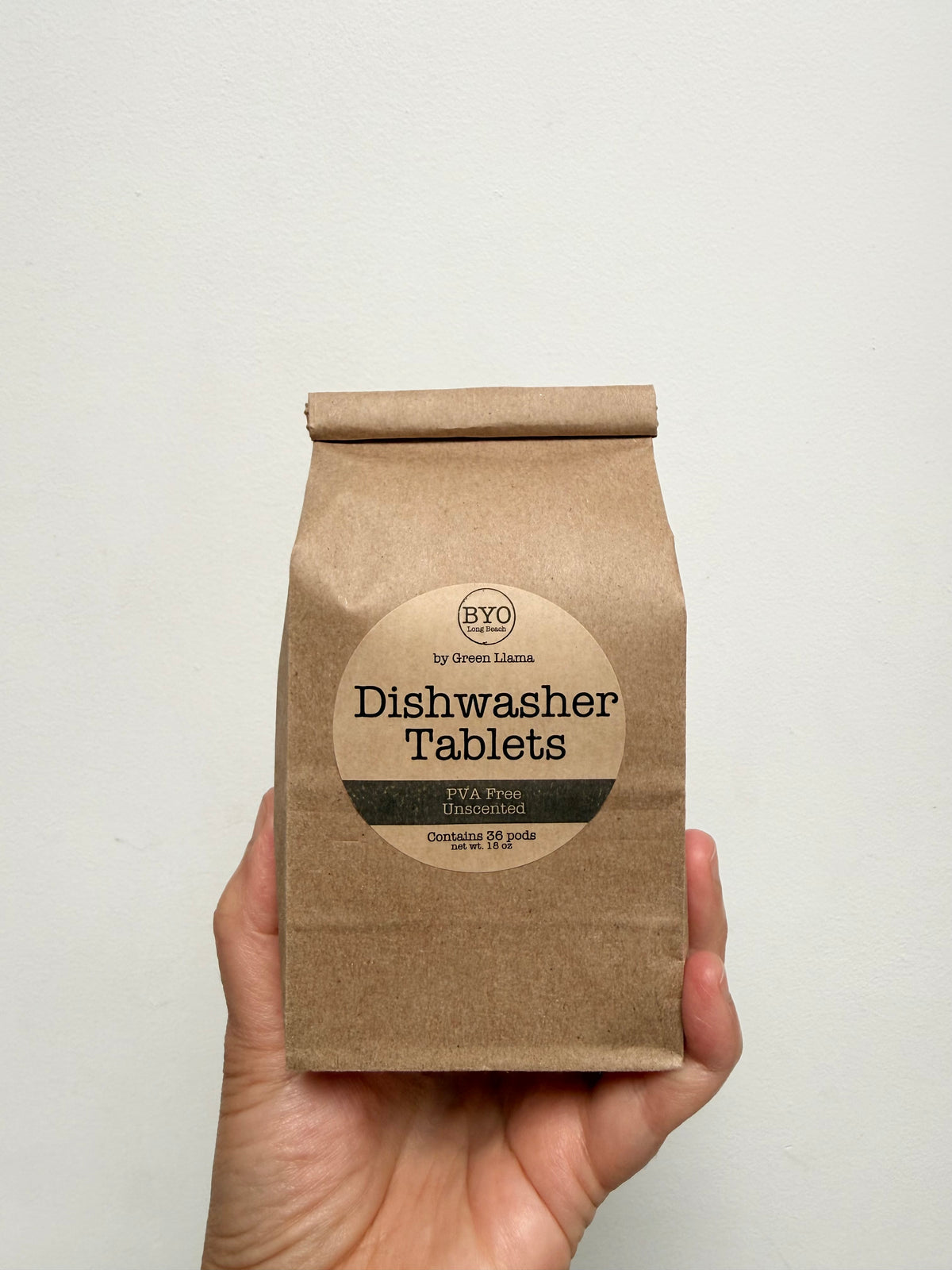 A hand holding a brown kraft paper bag of BYO Dishwasher Tablets against a plain off-white wall.