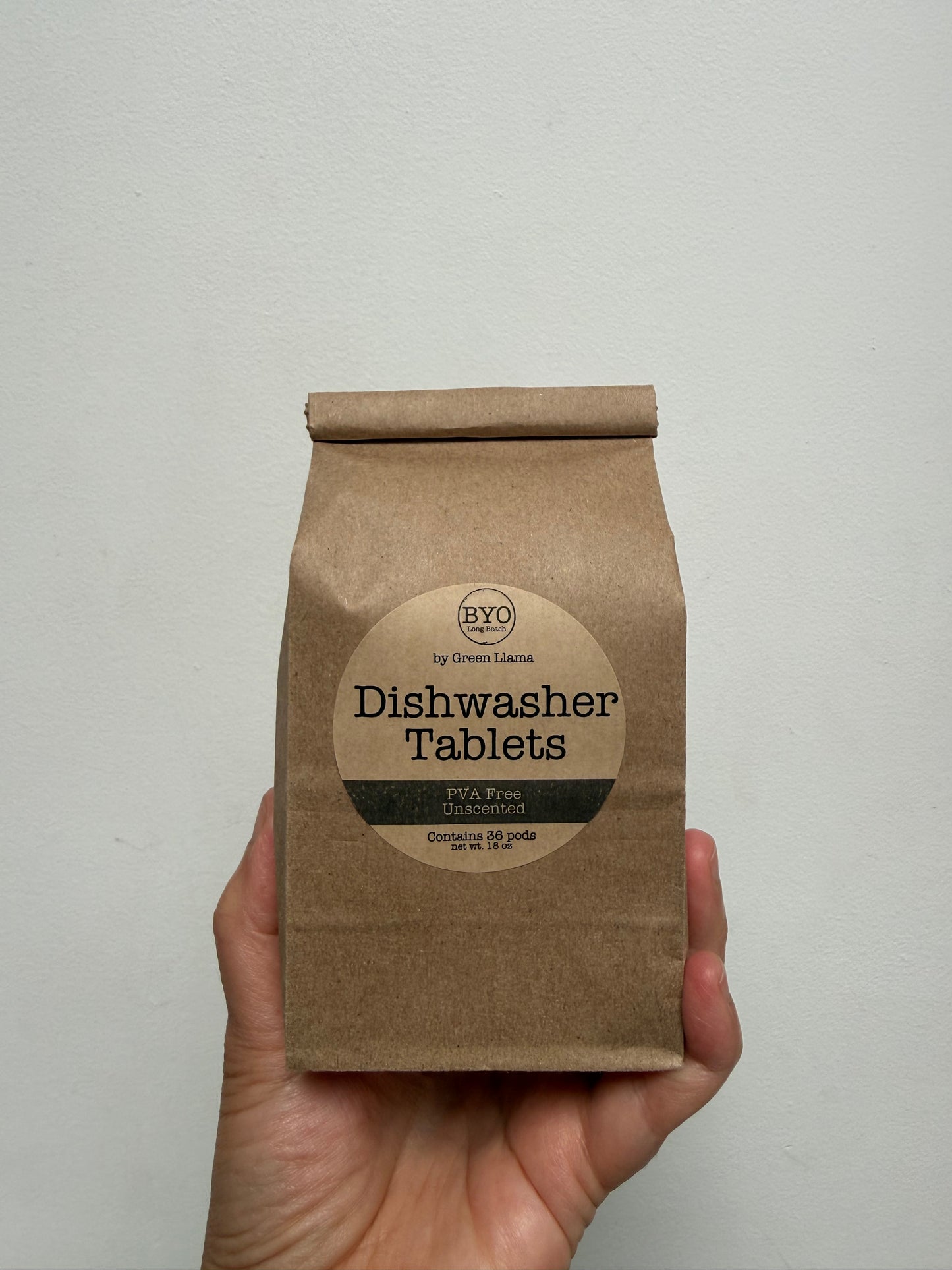 A hand holding a brown kraft paper bag of BYO Dishwasher Tablets against a plain off-white wall.