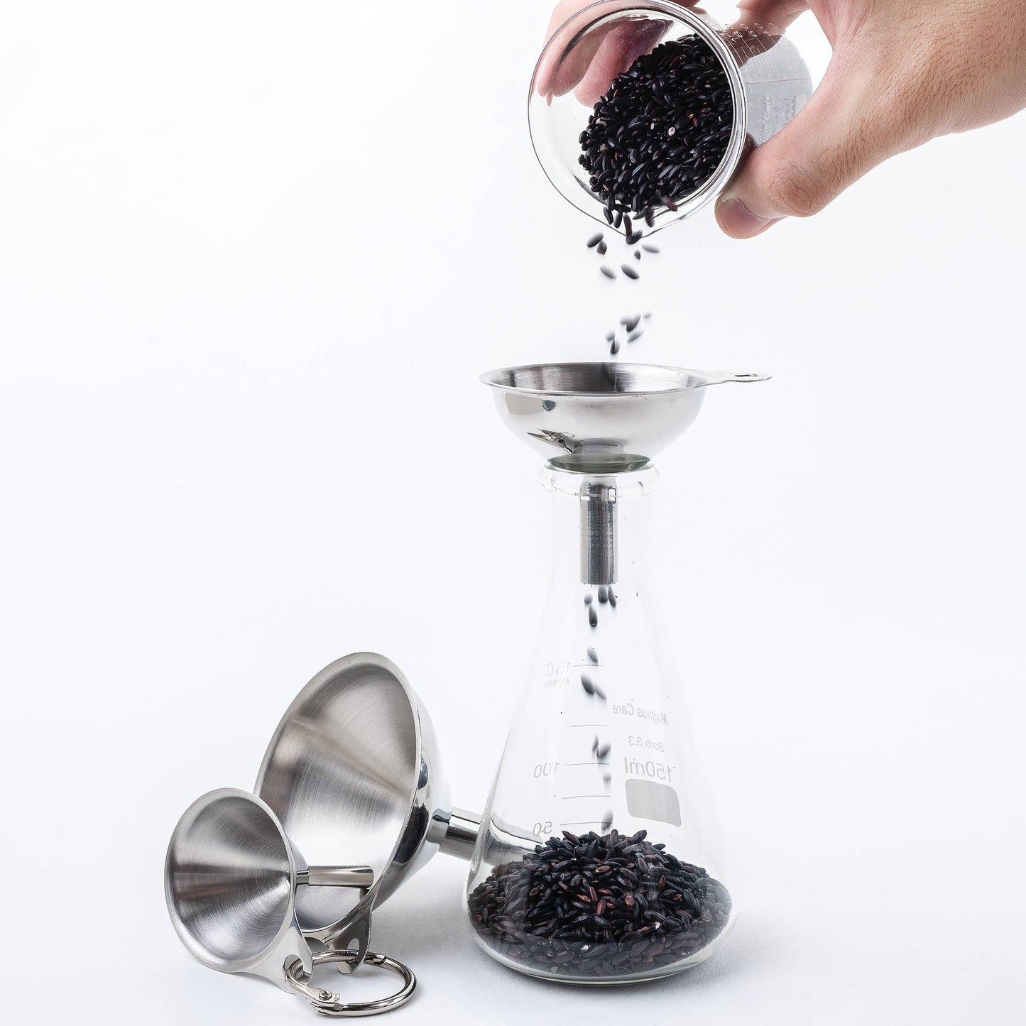 Hand pouring black seeds into a glass funnel with additional funnels on a white background