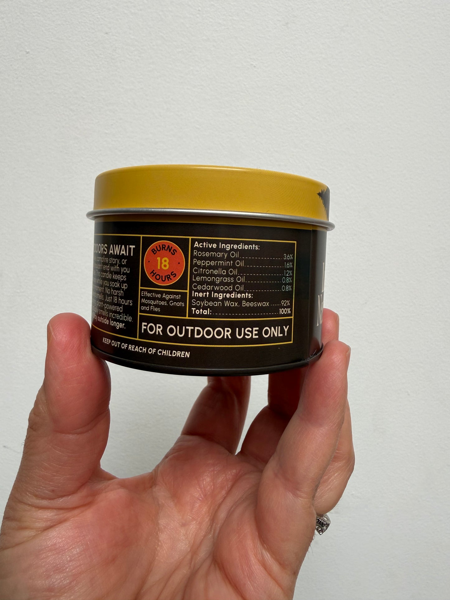 A hand holding a small cylindrical metal tin of Camp Fire outdoor candle against a plain off-white wall. The tin has a gold lid and a black label with white and orange text.
