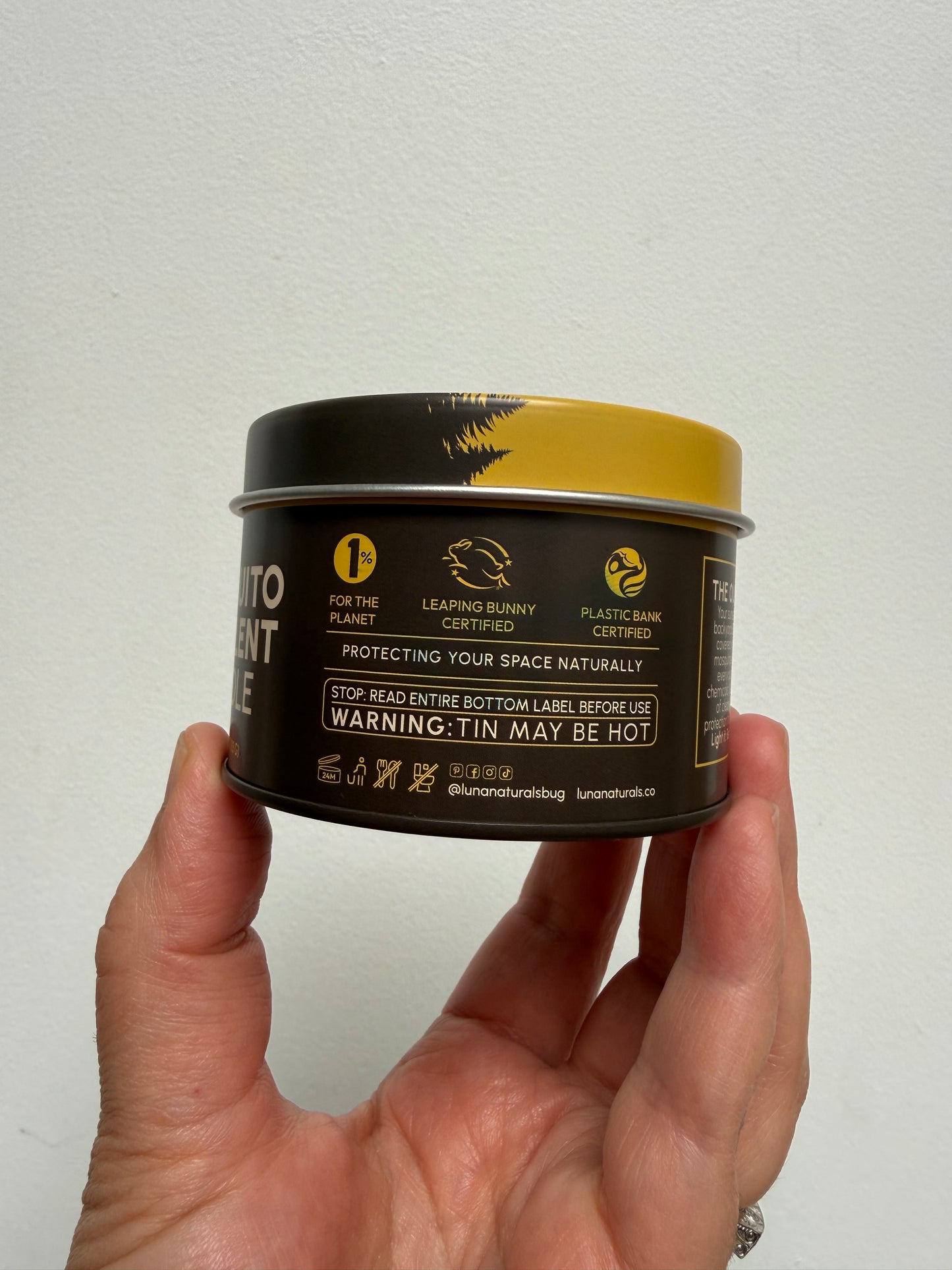 A hand holding a small cylindrical metal tin with a black label and gold lid, shown against a plain off-white wall. The label includes safety text and icons, including a prominent warning that says 'WARNING: TIN MAY BE HOT'.