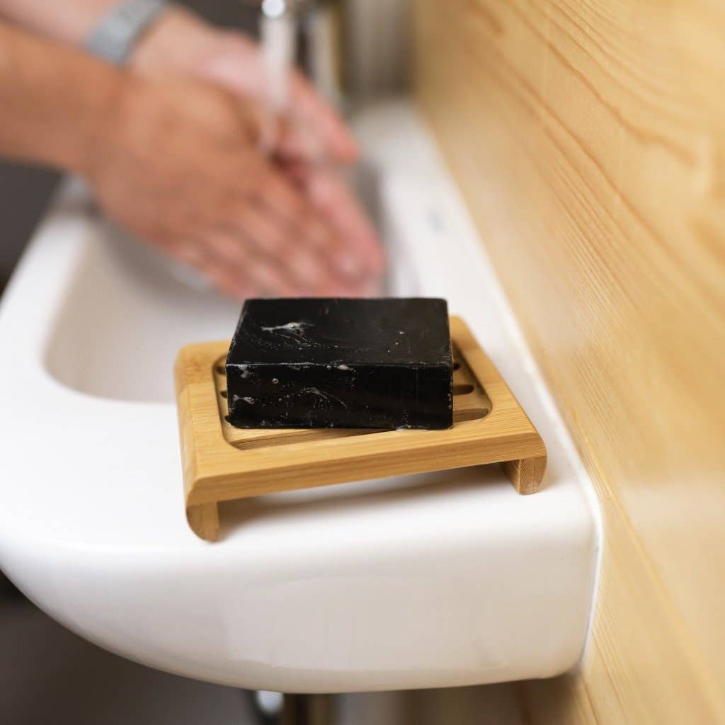Black soap bar on a wooden soap dish with hands washing in the background
