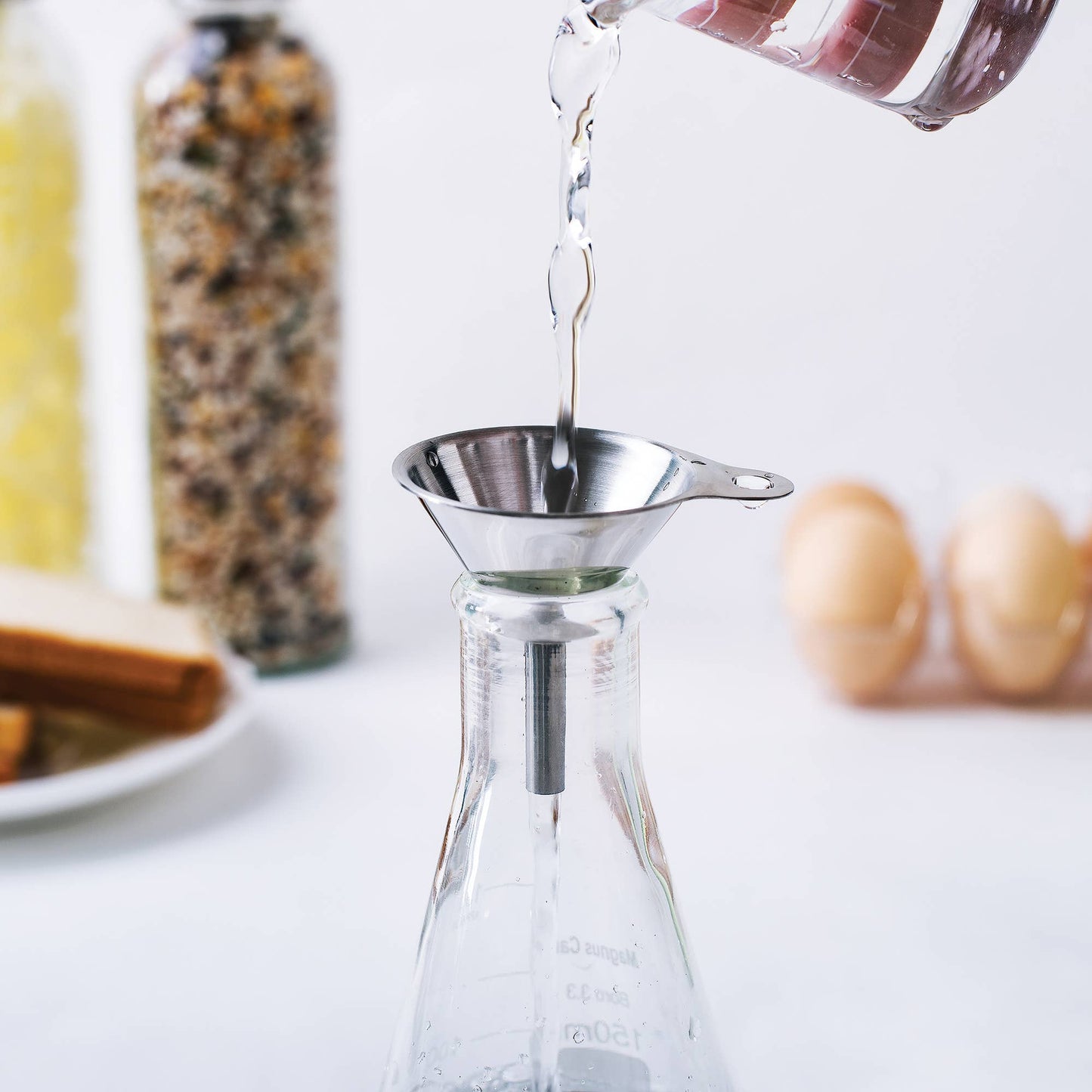 Pouring liquid from a glass container into a funnel over a clear carafe on a white surface.