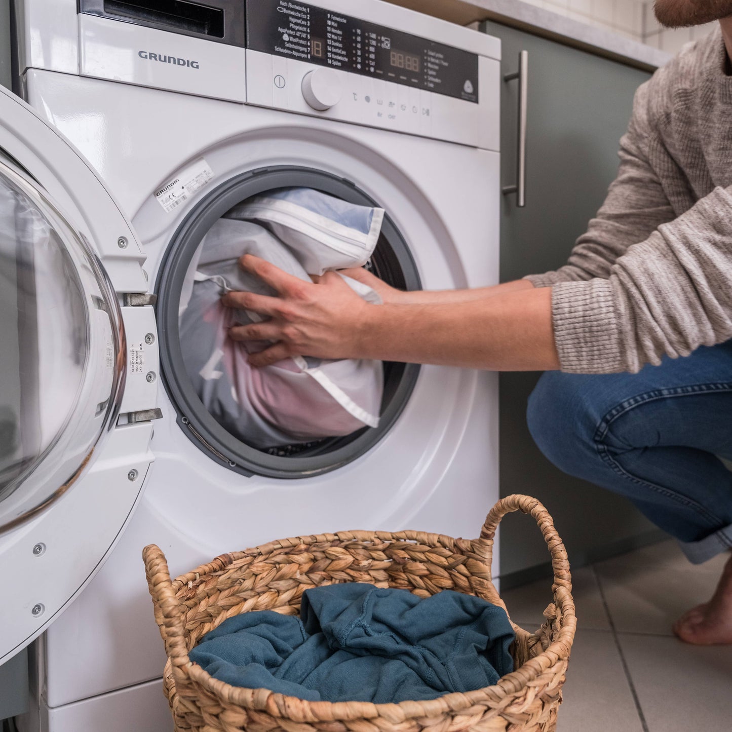 Person loading laundry into a washing machine with a basket of clothes nearby.