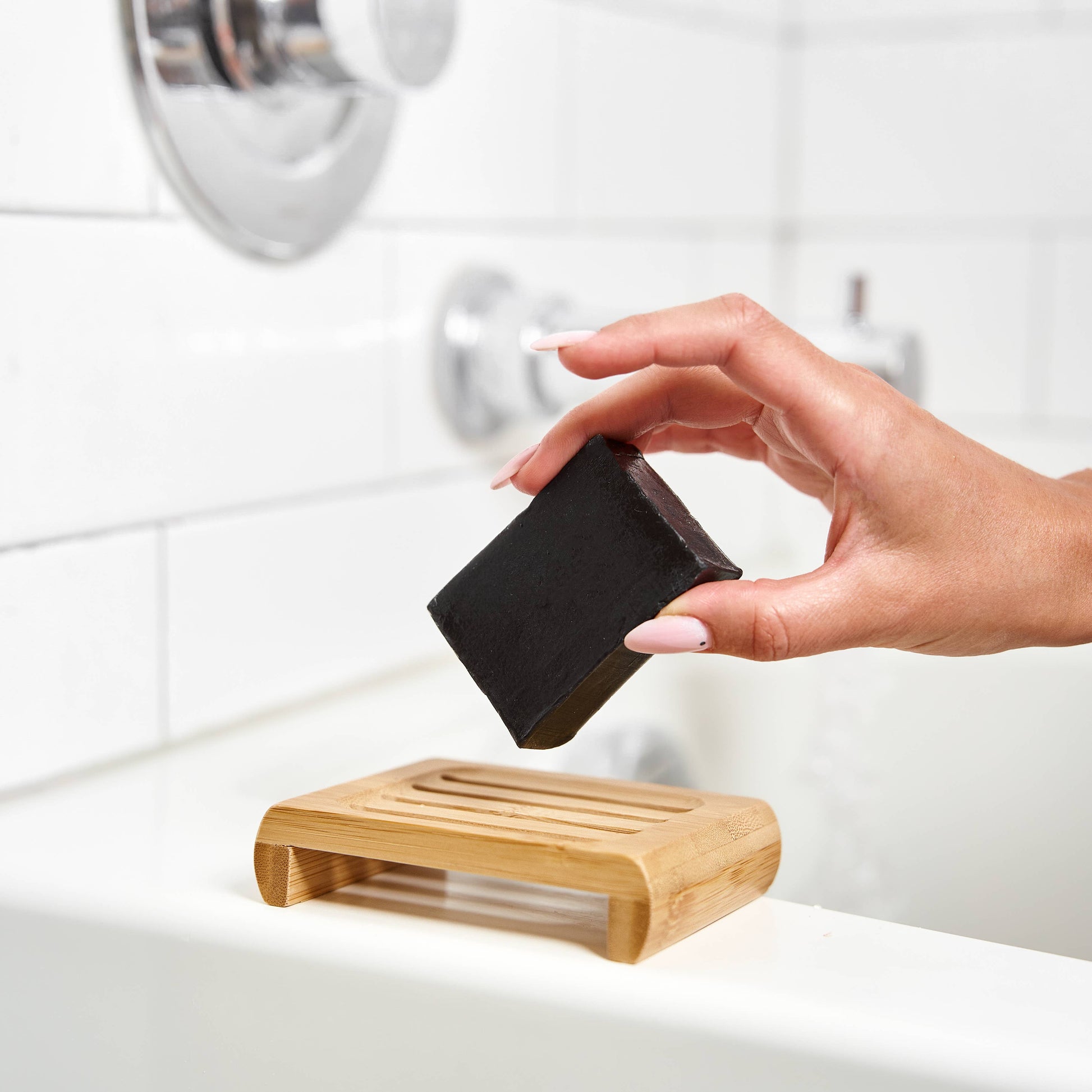 Hand holding a bar of soap over a wooden soap dish with a bathroom background.