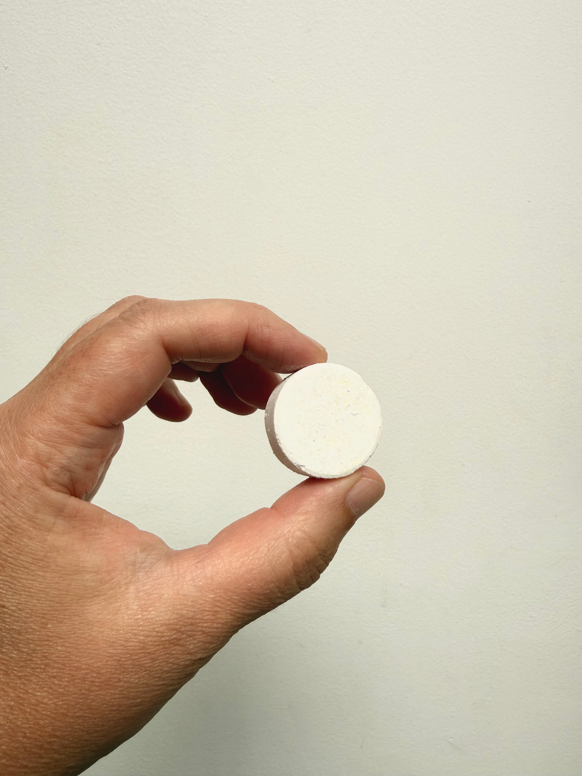 A hand holding a small, round, off-white tablet against a plain, light beige background.