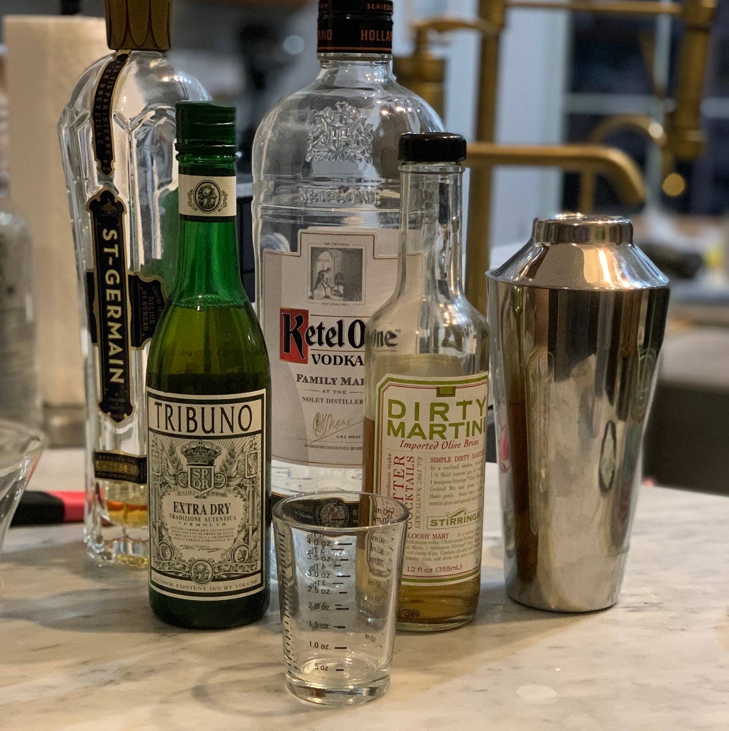 Alcohol bottles including St. Germain, Tribuno, Ketel One, and Dirty Martini on a bar counter with a cocktail shaker.