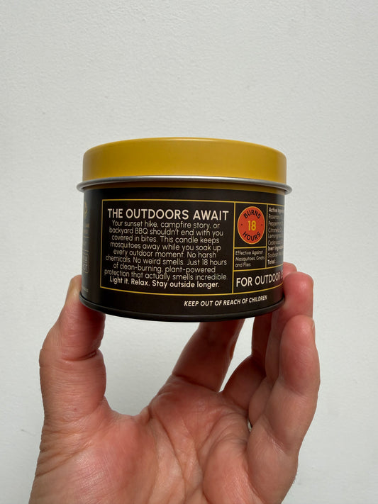 A hand holding a small round metal tin of The Outdoors Await against a plain off-white background.