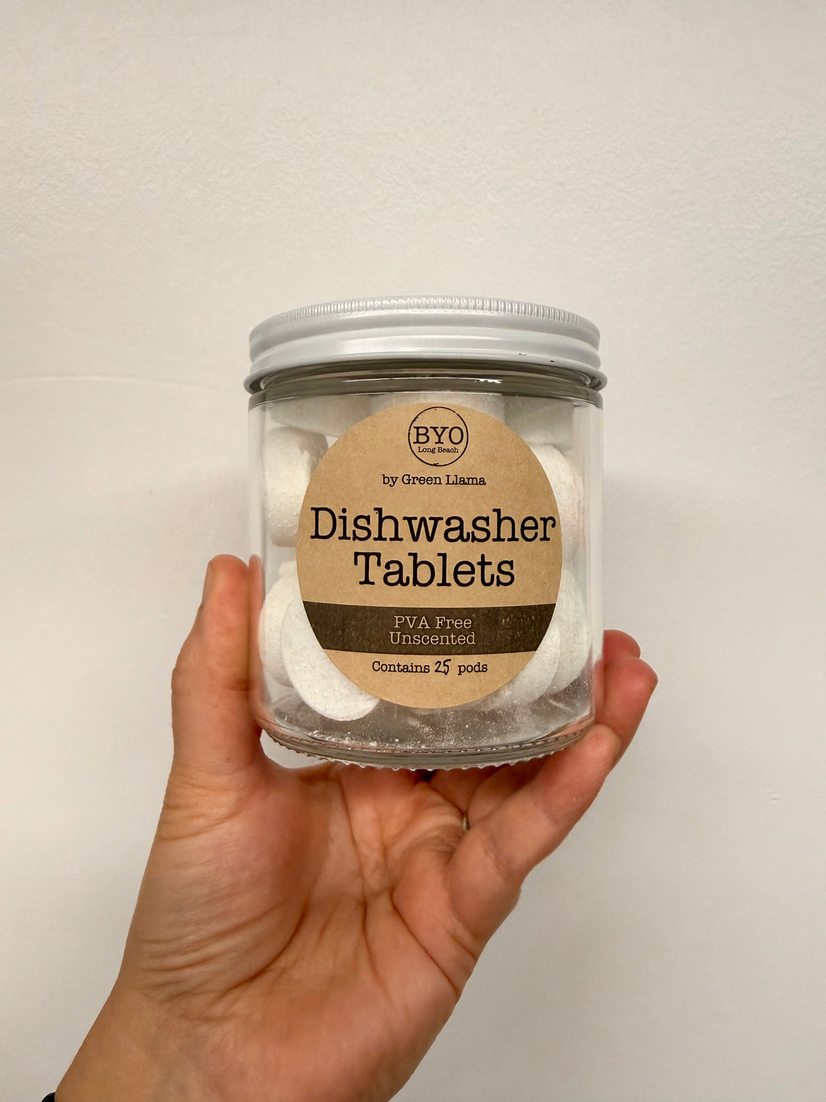 A hand holding a clear glass jar of BYO Dishwasher Tablets against a plain off-white wall. The jar has a silver metal lid and a round kraft paper label.