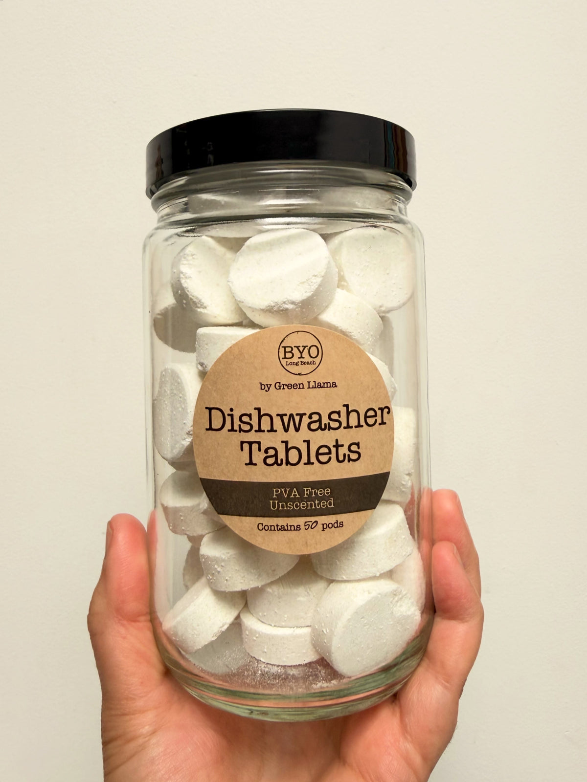 A hand holding a clear glass jar filled with white round dishwasher tablets, featuring a black lid and a brown paper label that reads 'BYO Dishwasher Tablets'.