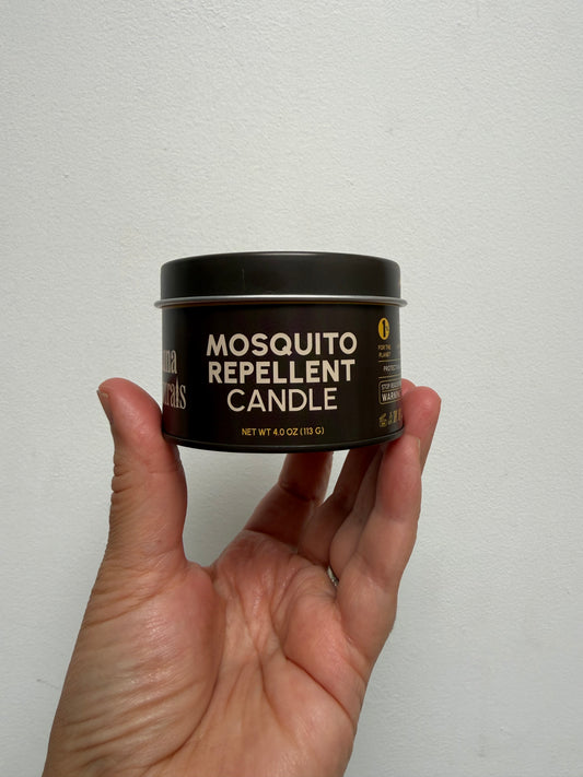 A hand holding a small black tin of mosquito repellent candle against a plain white wall.