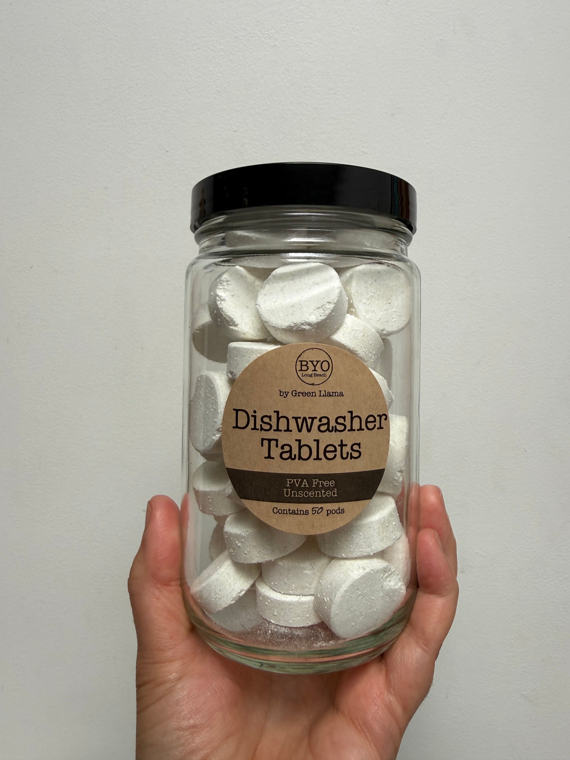 A hand holding a clear glass jar filled with white dishwasher tablets, featuring a black lid and a brown paper label that reads 'BYO Dishwasher Tablets'.