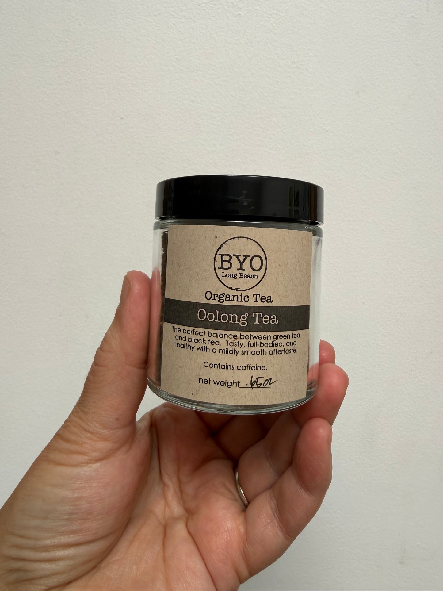 A hand holding a small clear jar of BYO Long Bean Organic Oolong Tea against a plain off-white wall.
