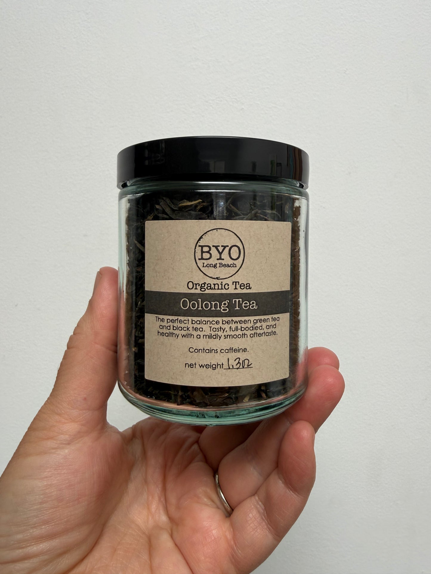 A hand holding a clear glass jar of BYO Organic Tea Oolong Tea against a plain white wall.