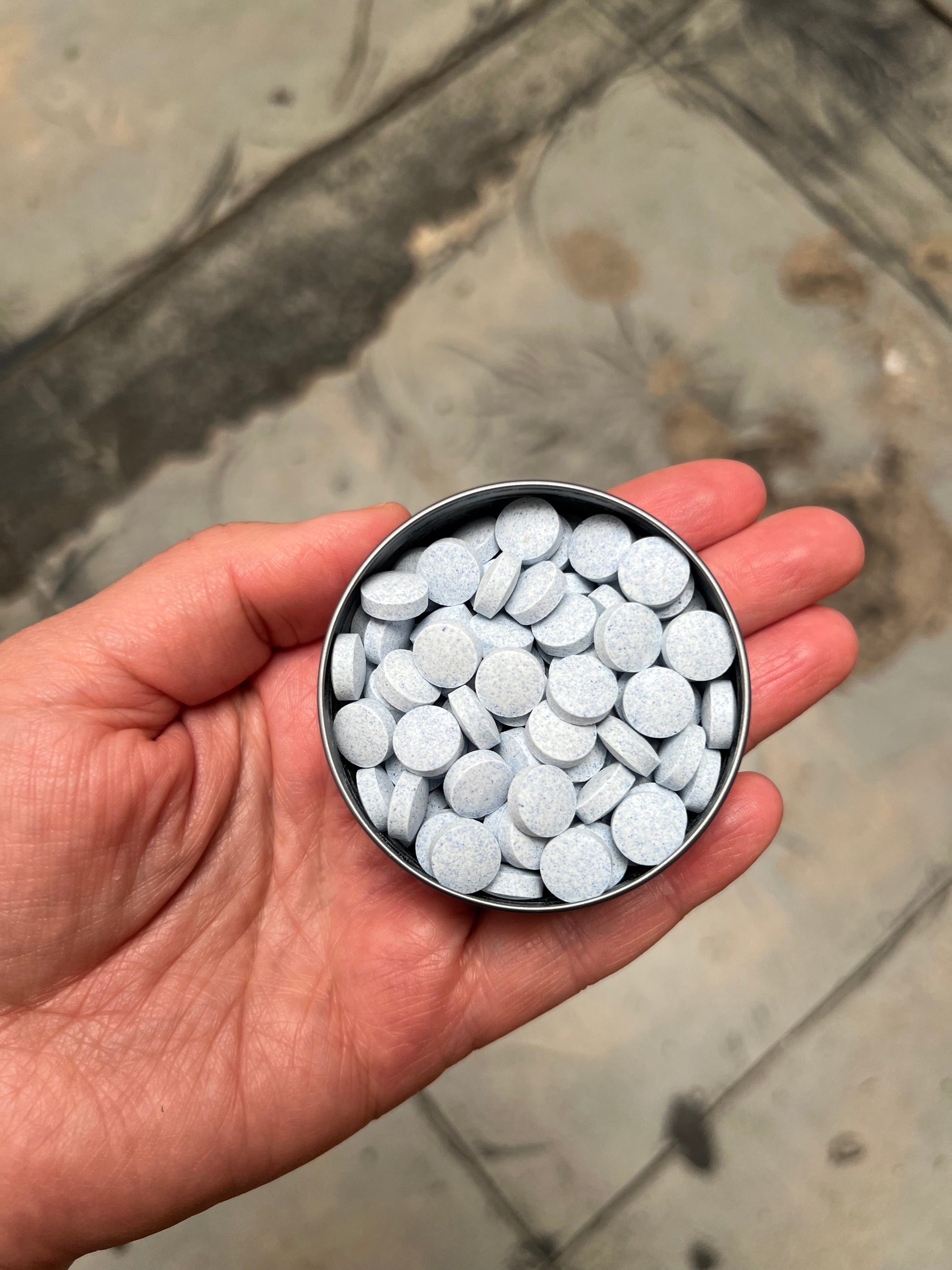 hand holding a metal tin with lid off showing light blue mouthwash tablets