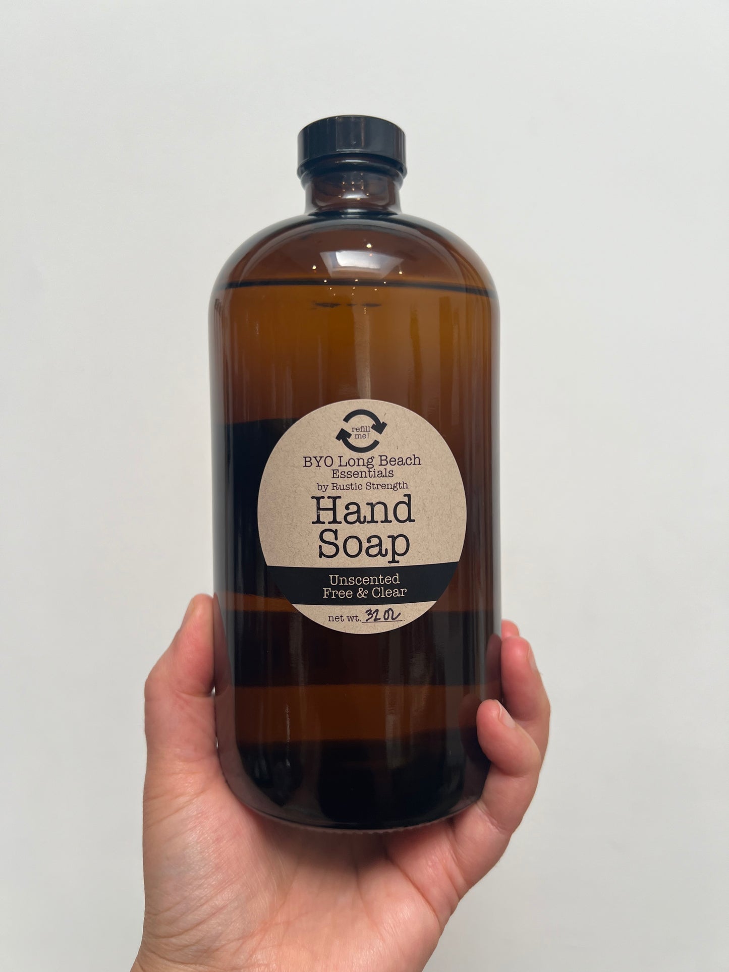 Hand holding a 32 ounce amber colored glass bottle with a black plastic cap. Circular label on bottle notes that it is hand soap.