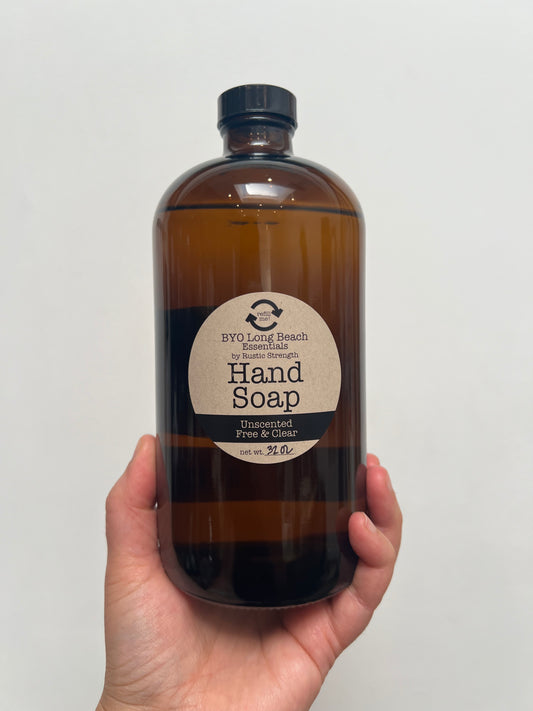 Hand holding a 32 ounce amber colored glass bottle with a black plastic cap. Circular label on bottle notes that it is hand soap.