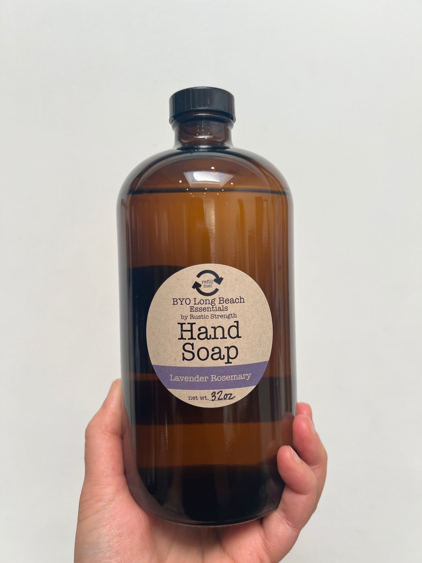 Hand holding a 32 ounce amber colored glass bottle with a black plastic cap. Circular label on bottle notes that it is hand soap.