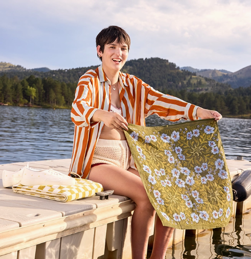 Woman sitting on a dock by a lake, holding a floral towel.