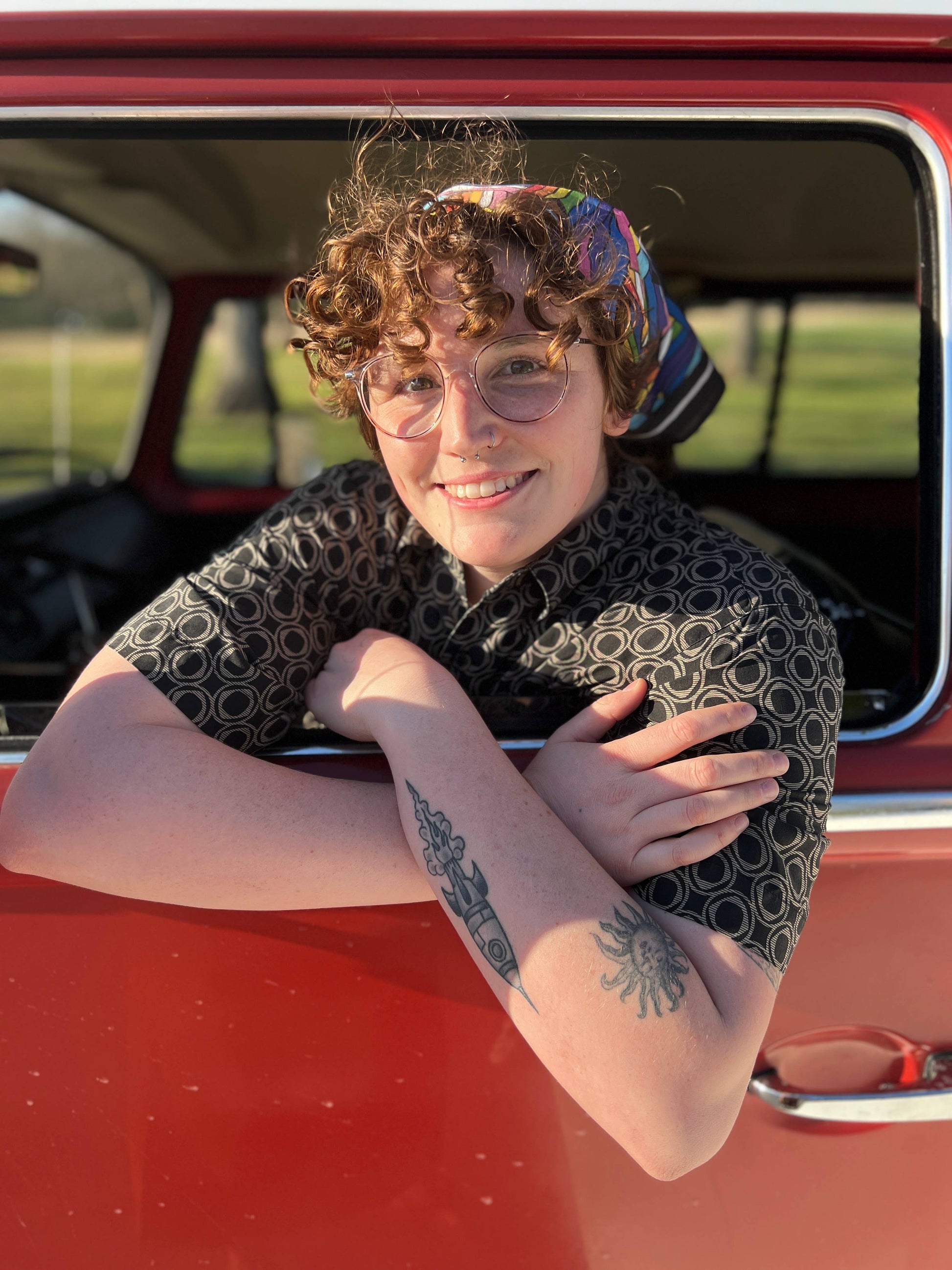 Person with tattoos leaning on a red vehicle, wearing a patterned shirt and headscarf.