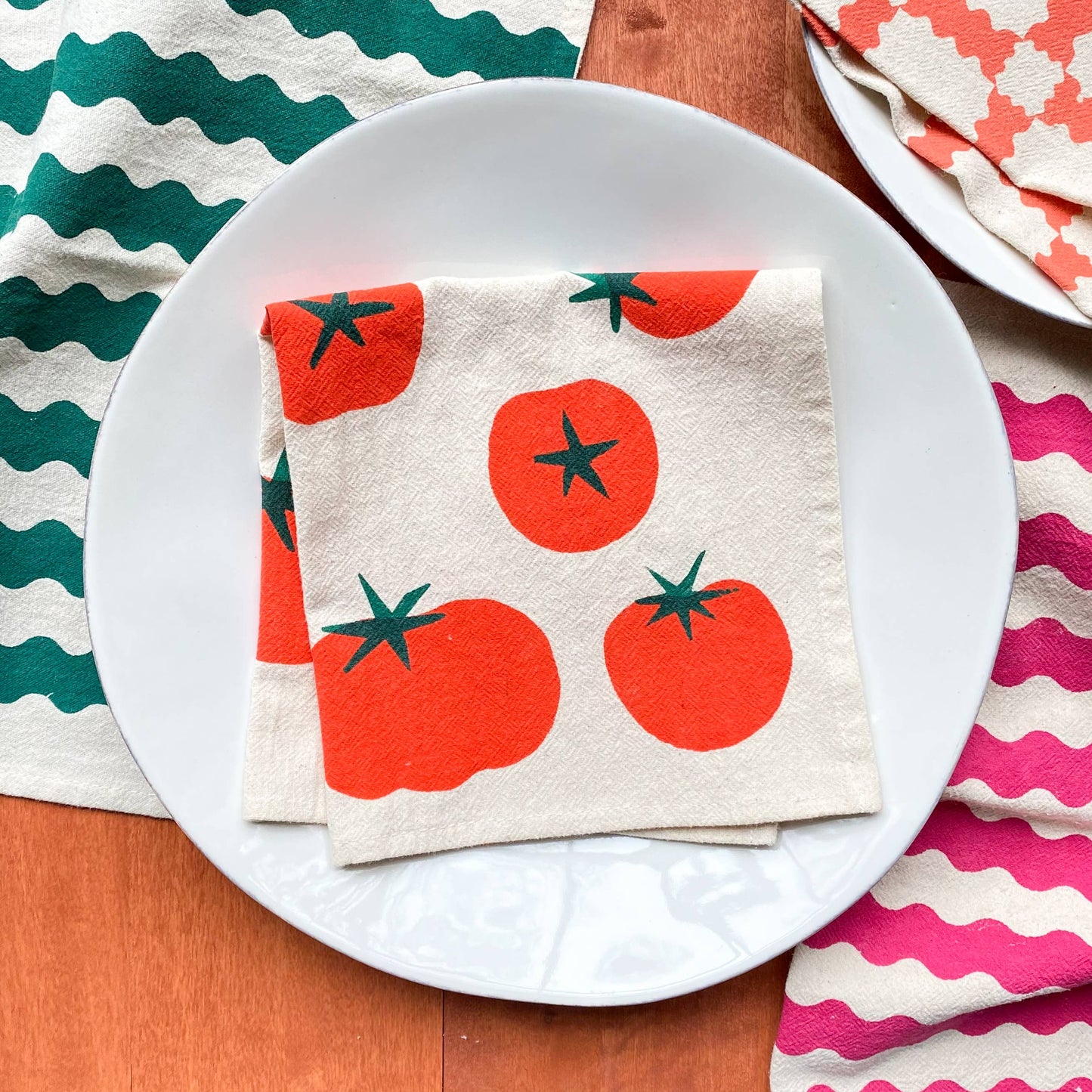 Napkins with tomato pattern on a white plate surrounded by colorful napkins.