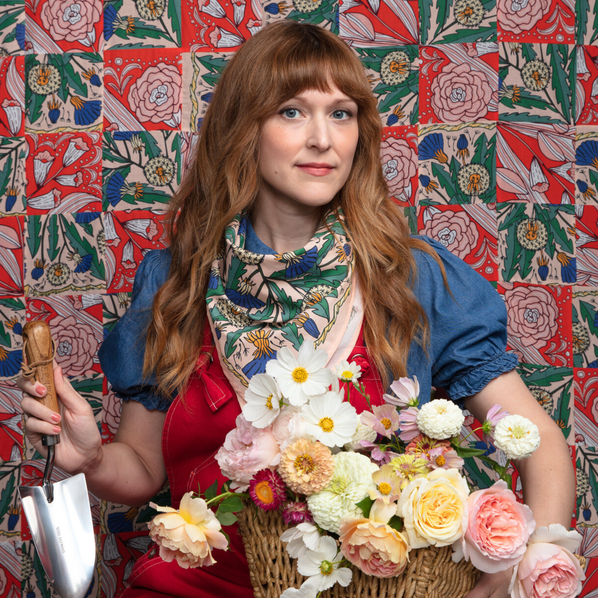 Woman holding a basket of flowers and a gardening tool against a colorful floral-patterned wall.