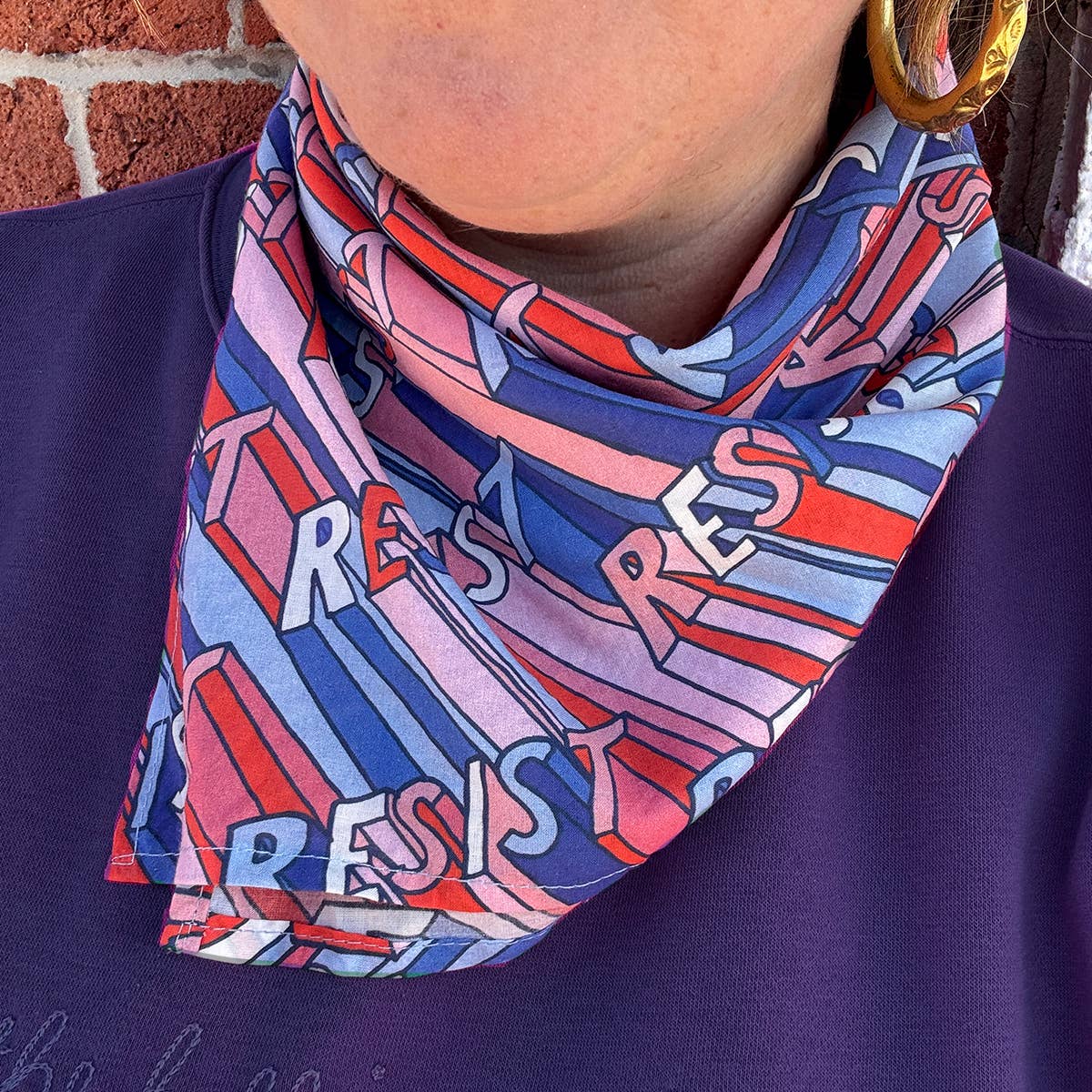 Person wearing a colorful scarf with abstract design and text, against a brick wall background.