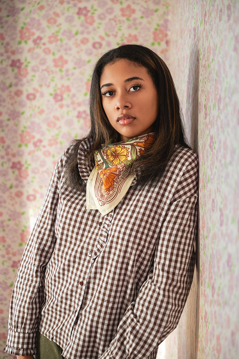 Woman wearing a checkered shirt with a patterned scarf against a floral wallpaper background